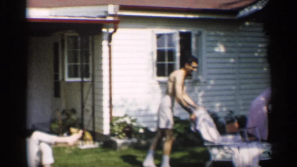 A man appears to move or adjust a lounge chair near the side of a white house during a sunny 1959 Indiana backyard scene.