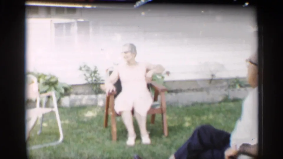 A woman in a light dress sits outdoors on a wooden chair against a white house wall in 1959 Indiana home-movie footage.