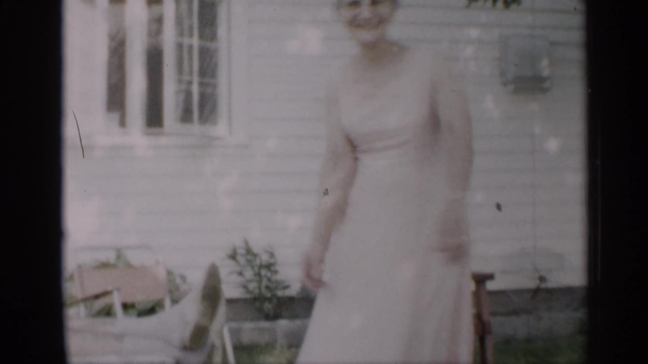 Woman in a pale sleeveless dress moves in a yard beside a white-sided house.