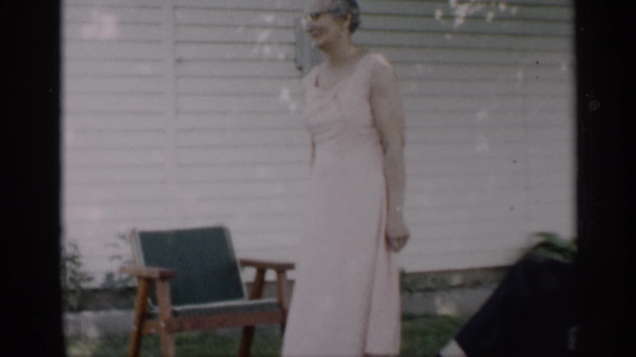A woman wearing glasses and a pale sleeveless dress smiles outside an Indiana house in 1959 home-movie footage.