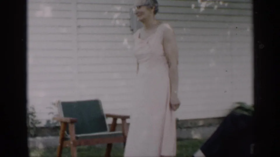 A woman wearing glasses and a pale sleeveless dress smiles outside an Indiana house in 1959 home-movie footage.