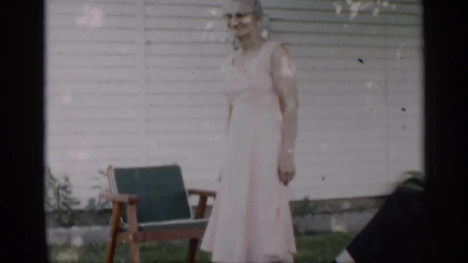 The 1959 Indiana yard scene shows the woman paused beside a wooden chair, with house siding and grass behind her.