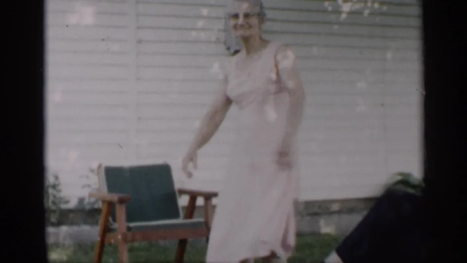 A woman in a pale dress steps across the yard in Indiana, with a wooden chair and white house siding visible behind her.