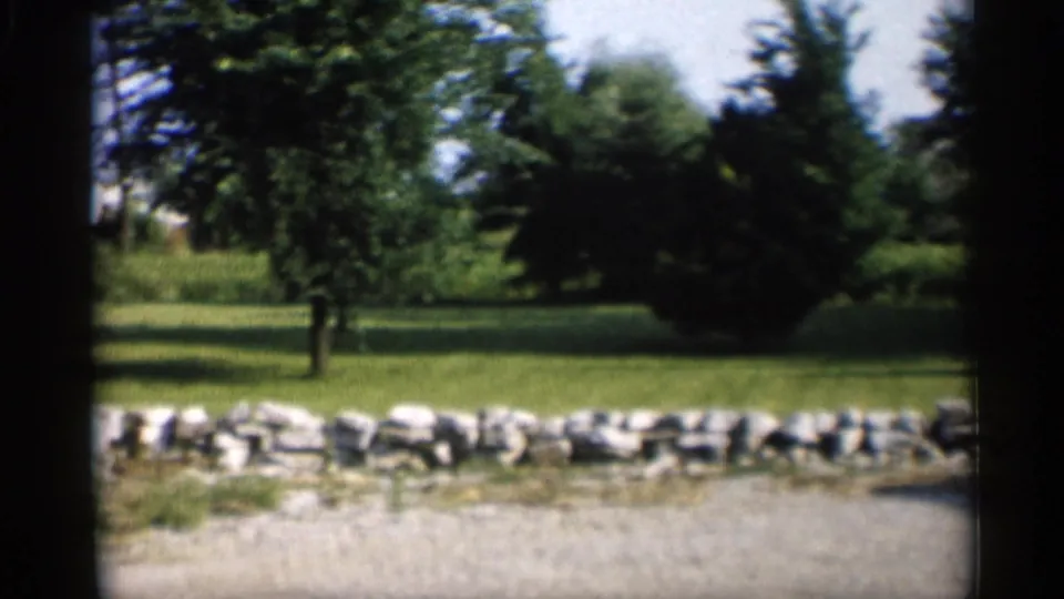 1959 Indiana home-movie view of a low rock border running along a lawn with trees, shrubs, and a gravel driveway in front.