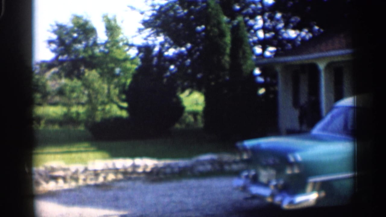 Turquoise 1950s car parked beside a yellow house and lawn.
