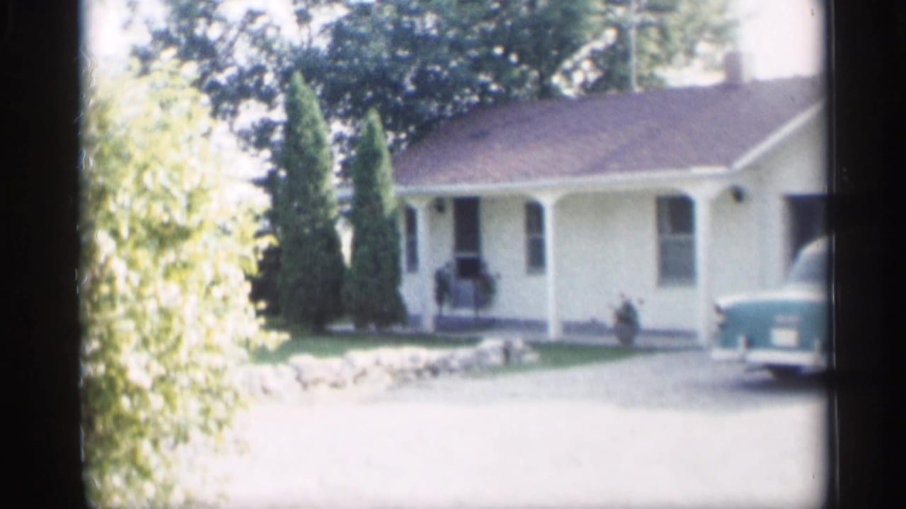 Yellow house with shrubs and part of a turquoise car at the driveway edge.
