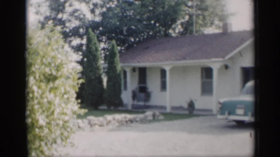 1959 Indiana 8mm frame showing the front of a yellow house with tall shrubs, low stone wall, and turquoise car rear.