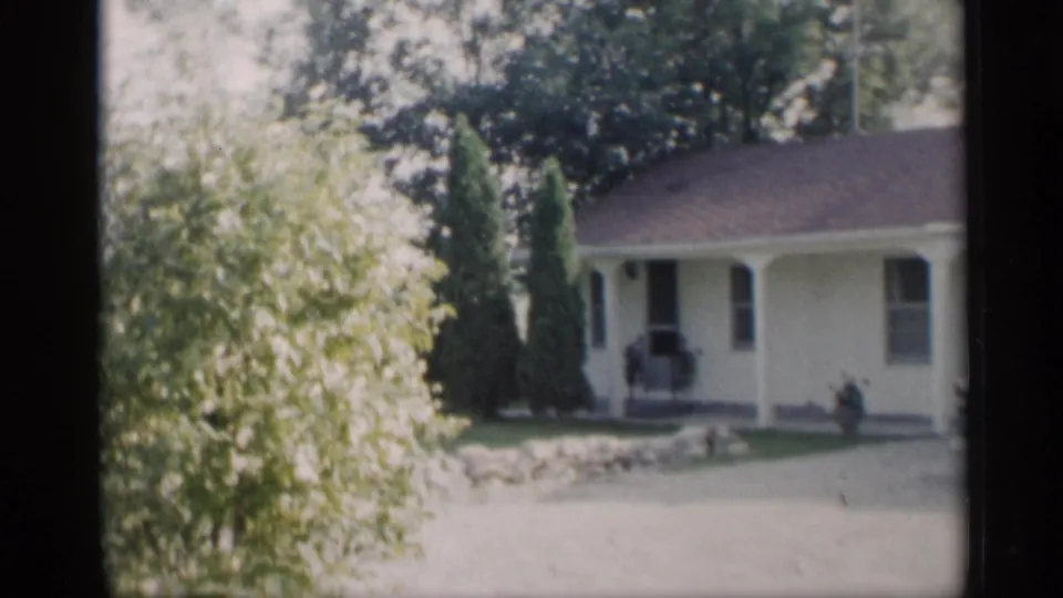 1959 Indiana home-movie frame of a yellow suburban house, porch, shrubs, lawn, and bright driveway.