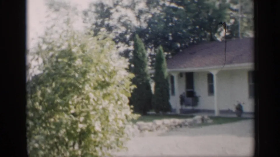 1959 Indiana 8mm frame showing a white suburban ranch house with porch, shrubs, lawn, and trees in soft home-movie focus.