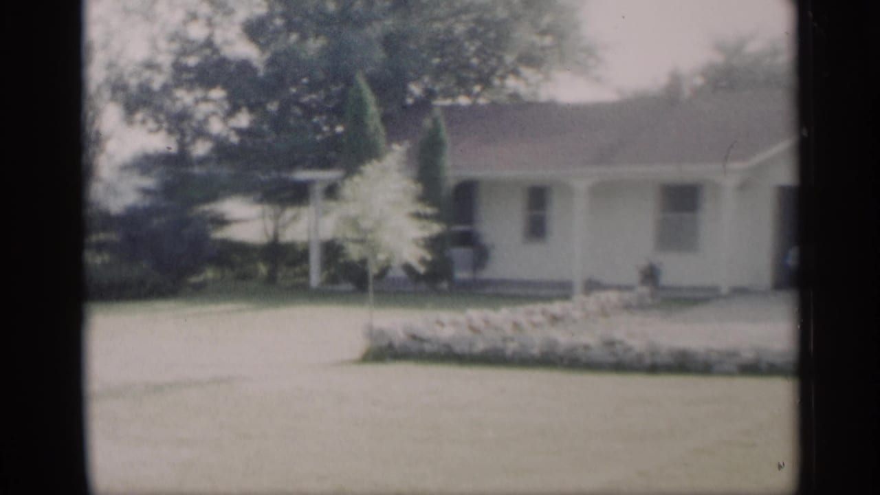 1959 Indiana home-movie view of a ranch house exterior with a wide lawn, small ornamental tree, stone border, and covered porch.