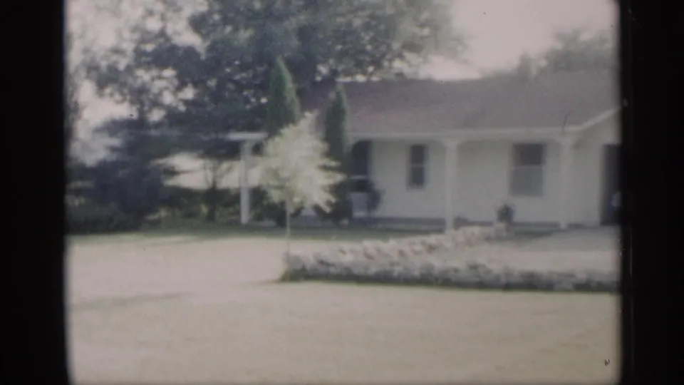 1959 Indiana home-movie view of a ranch house exterior with a wide lawn, small ornamental tree, stone border, and covered porch.