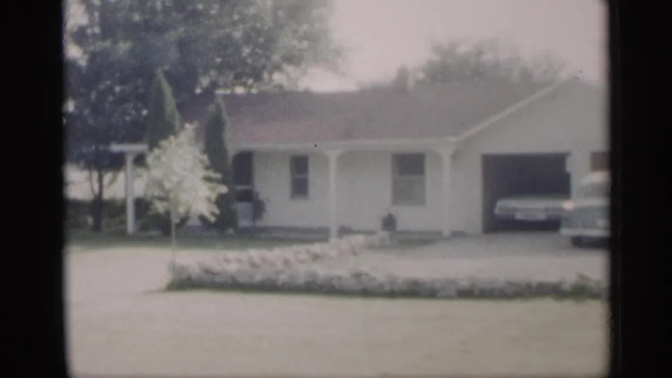 1959 Indiana 8mm frame showing the ranch house driveway, garage opening, lawn border, porch, and a parked car partly visible.