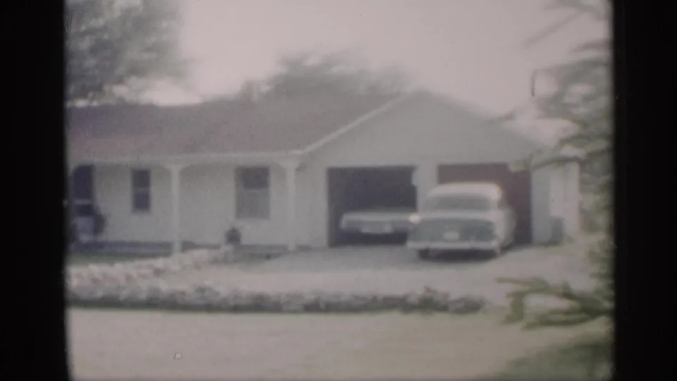 1959 Indiana home-movie frame of a ranch house with garage, driveway, two parked cars, front lawn, and stone-edged landscaping.