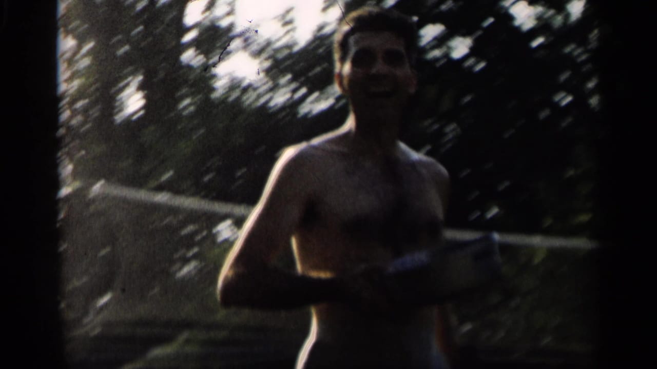 Shirtless man holding a badminton racket near a net on a shaded lawn.
