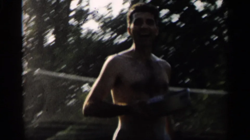 In 1959 Indiana home-movie footage, a shirtless adult man stands by a backyard badminton net with trees behind him.