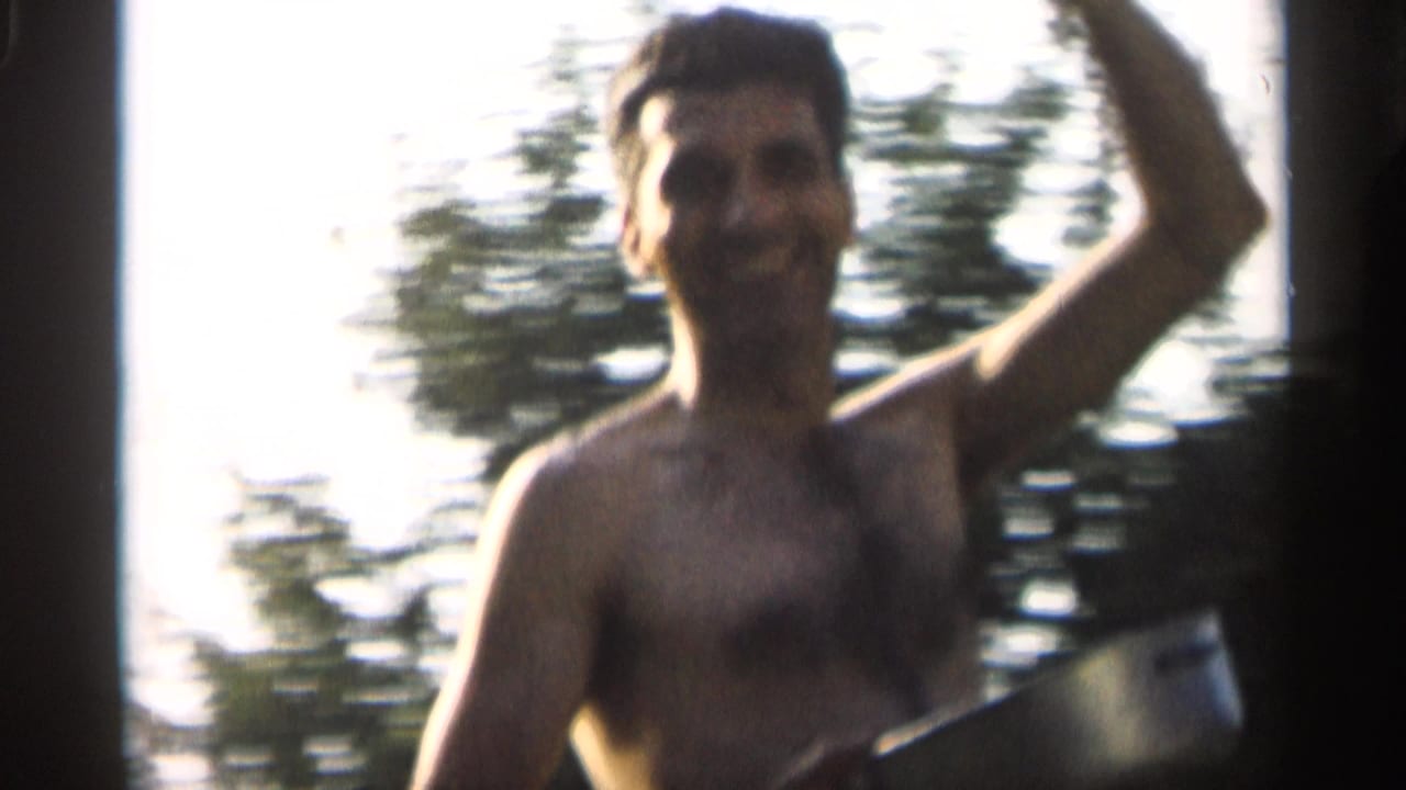 Shirtless man smiles and raises one arm while holding a badminton racket outdoors.