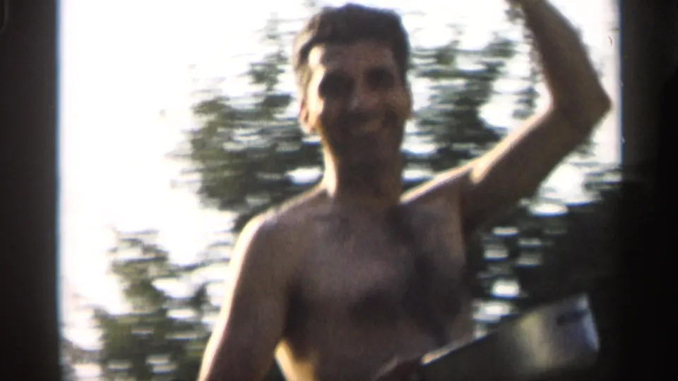 A shirtless adult man smiles toward the camera during a backyard badminton game in Indiana archival 8mm footage.