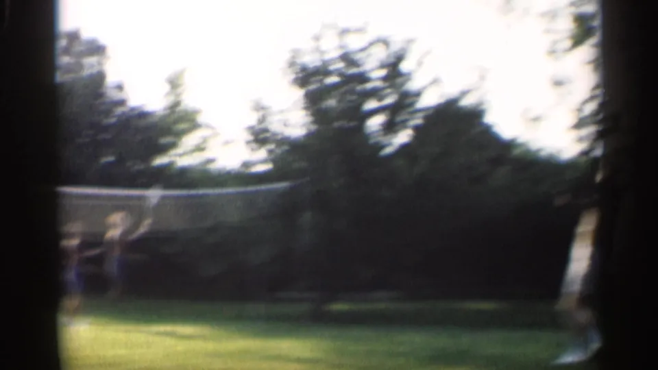 A distant player appears near the badminton net on a sunlit Indiana lawn, with trees and heavy film blur in the frame.
