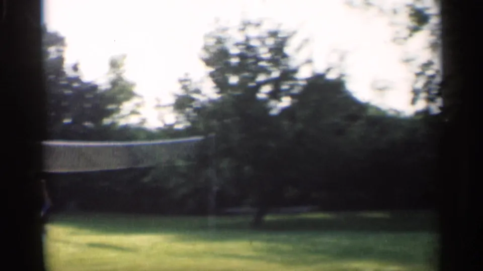 The camera frames the backyard badminton net and open lawn in 1959 Indiana home-movie footage, with trees beyond.