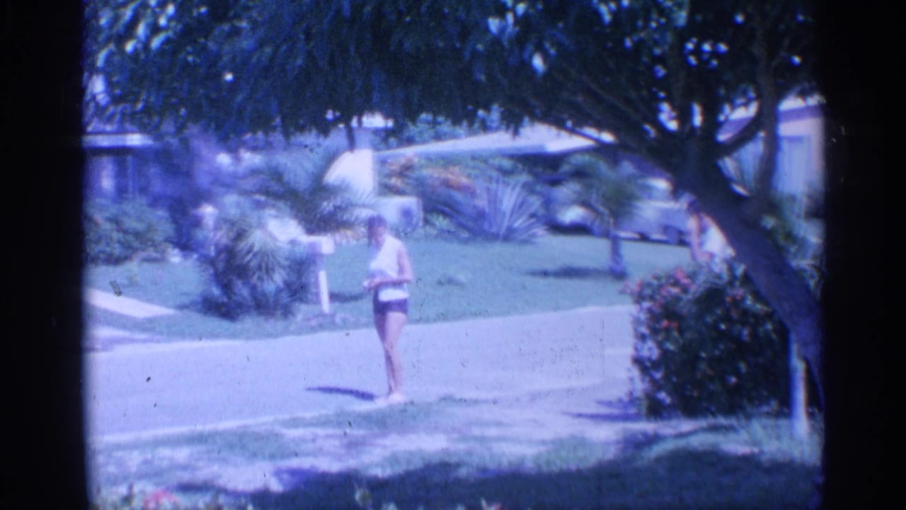 In supported 1959 Indiana home-movie footage, a woman walks near a suburban street with lawns, shrubs, trees, and house exteriors behind her.