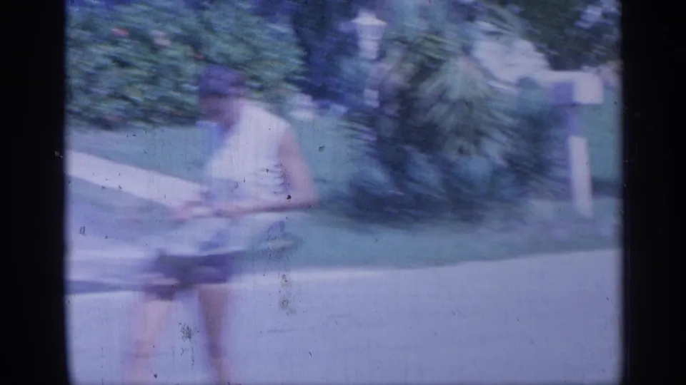 A woman in light clothing moves through a soft, blue-cast 8mm frame beside shrubs, lawn, and a pale roadside mailbox.