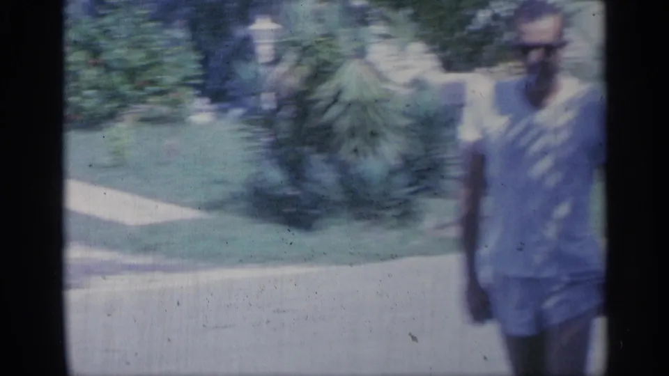 A man in a light shirt and shorts walks close to the camera, with lawns, shrubs, a sidewalk edge, and house details in the background.