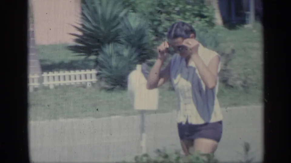 A woman in a light sleeveless top and dark shorts adjusts sunglasses or hair while walking near a lawn, mailbox, shrubs, and house exterior.