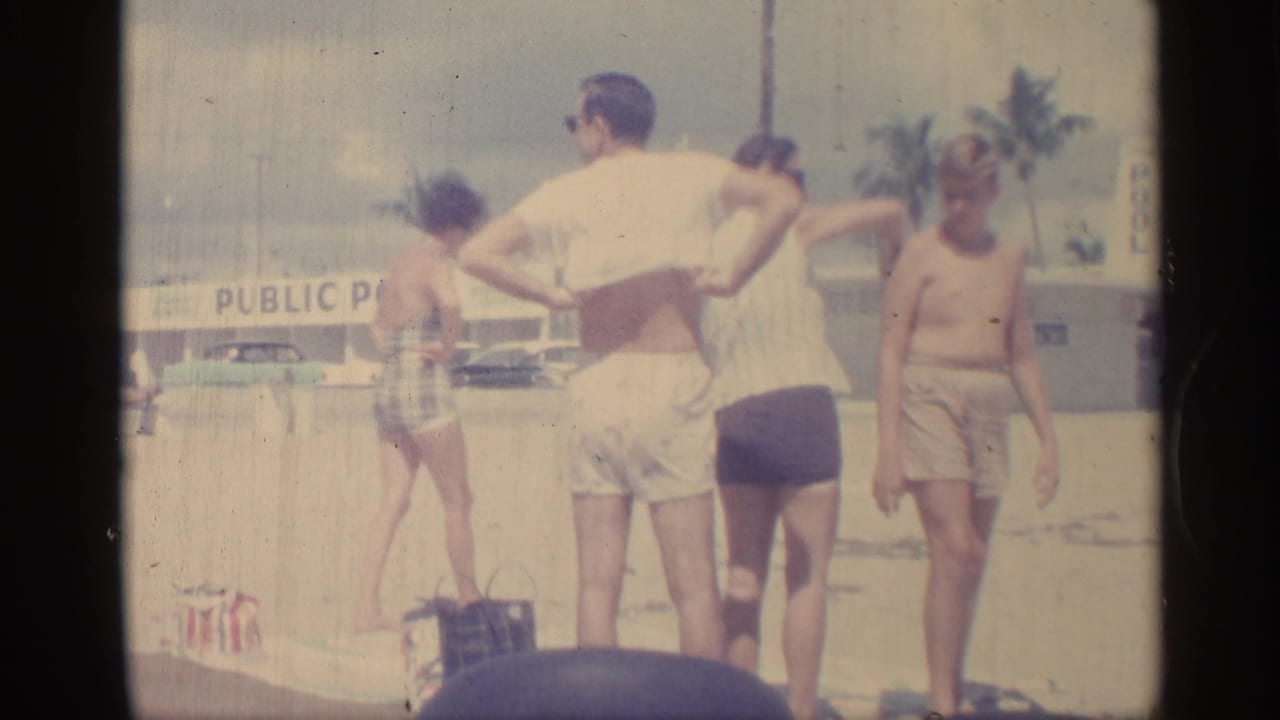 1959 Indiana home-movie frame of adults in sunglasses and swimwear standing near a PUBLIC POOL sign on a bright sandy poolside area.