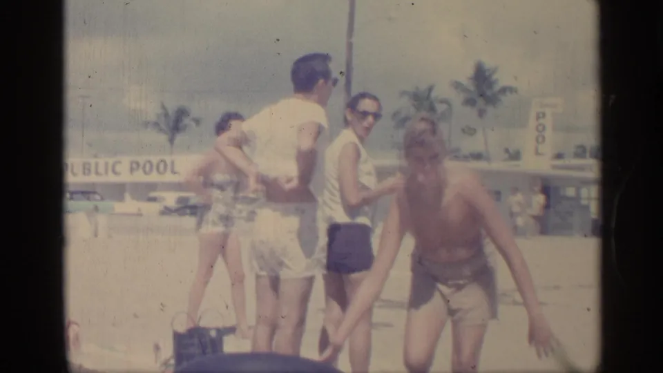 1959 Indiana poolside frame showing adults in period swimwear and sunglasses, with one man bending forward near the sandy area.