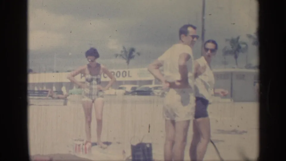 1959 Indiana home-movie frame with three adults in swimwear and sunglasses standing near a PUBLIC POOL sign and beach bags.
