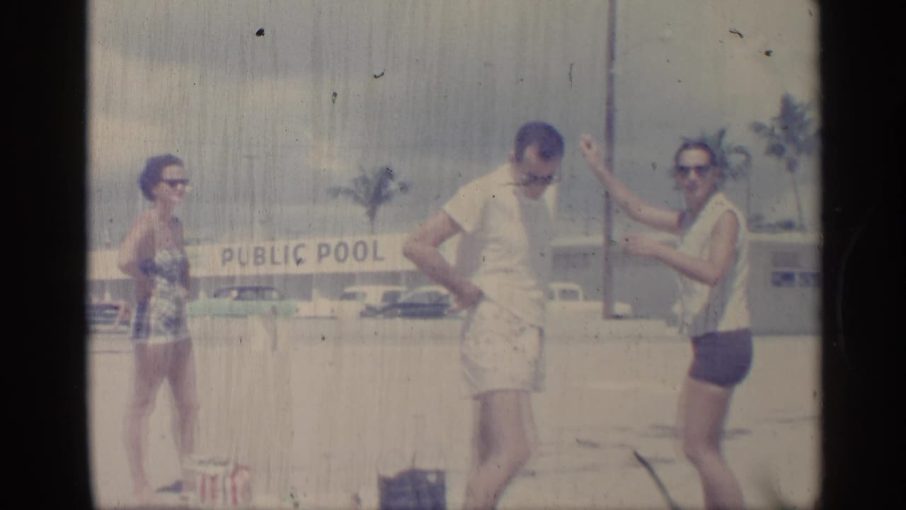 In this 1959 Indiana home-movie frame, adults in beachwear stand near a PUBLIC POOL sign with period cars and palm trees in the background.