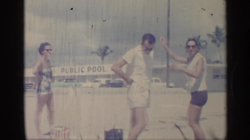 In this 1959 Indiana home-movie frame, adults in beachwear stand near a PUBLIC POOL sign with period cars and palm trees in the background.