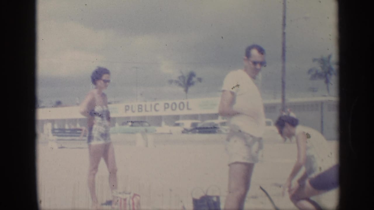 Adults stand and bend near beach bags in front of a public pool sign.