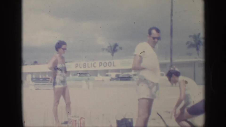Adults in 1959 beachwear stand beside bags near a PUBLIC POOL sign, with cars and palm trees visible beyond the bright sandy area.