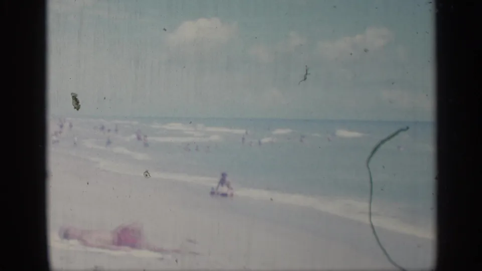 The shoreline frame shows distant swimmers, a seated figure near the water, and a sunbather on the sand in faded 8mm color.