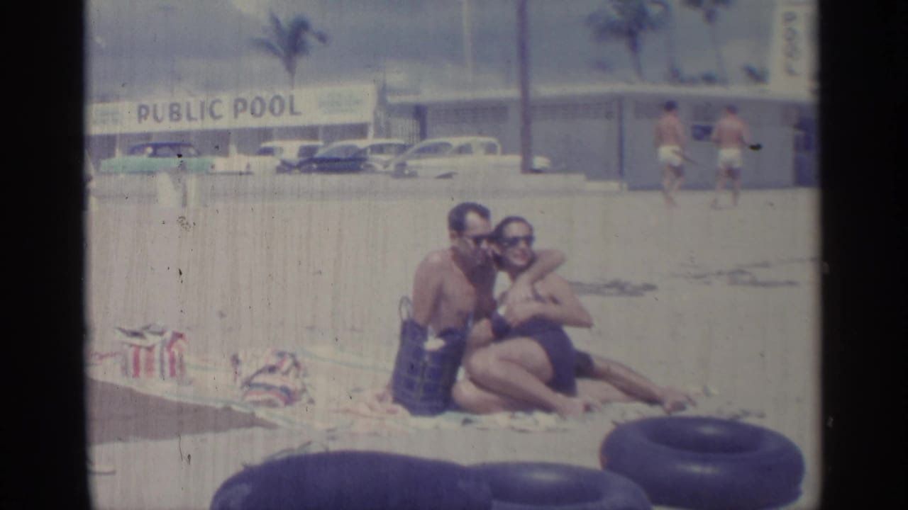A couple sits close together on a sandy beach near dark inner tubes and a public pool sign.