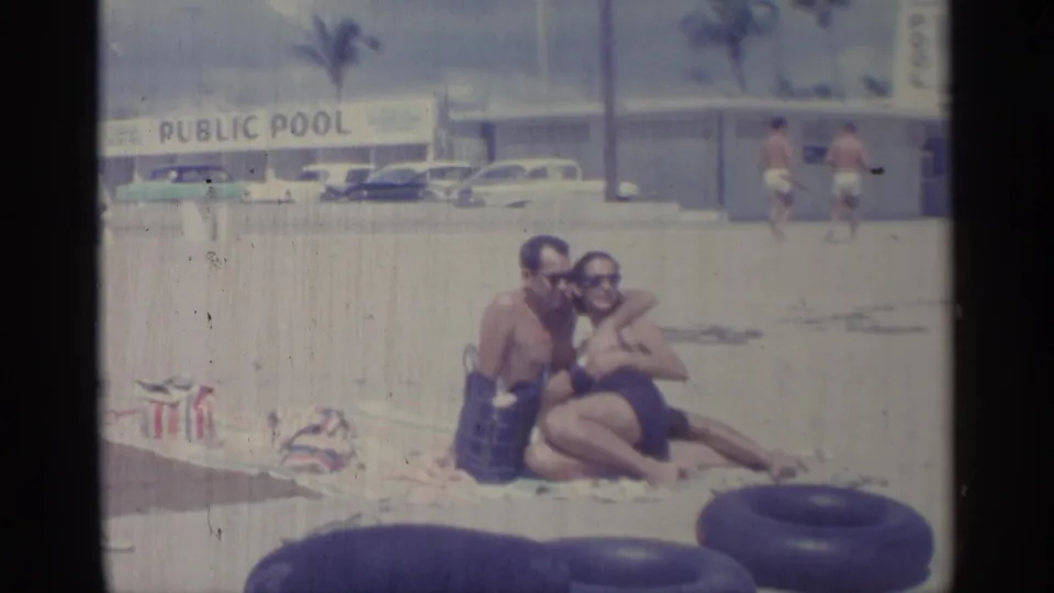 1959 Indiana home-movie frame of two adults in swimsuits sitting on sand near rubber inner tubes, with a public pool sign and parked cars behind them.