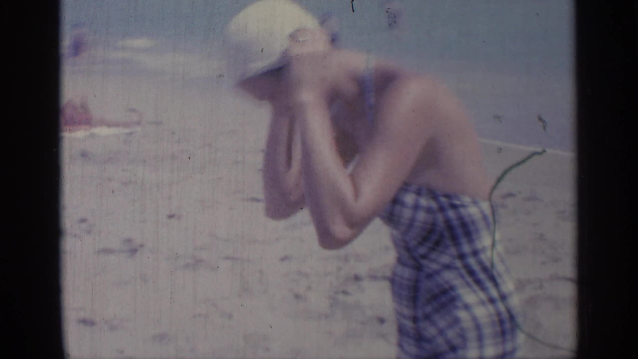 A woman in a plaid swimsuit and white swim cap adjusts her cap on the beach.