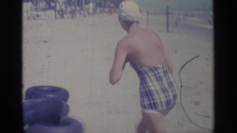 1959 Indiana home-movie frame of a woman seen from behind in a plaid swimsuit and swim cap walking across sand beside dark inner tubes.
