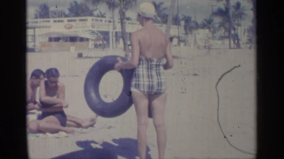 1959 Indiana beach frame of a woman in a plaid swimsuit holding a dark inner tube, with seated adults, palm-like trees, and public pool signage in the background.