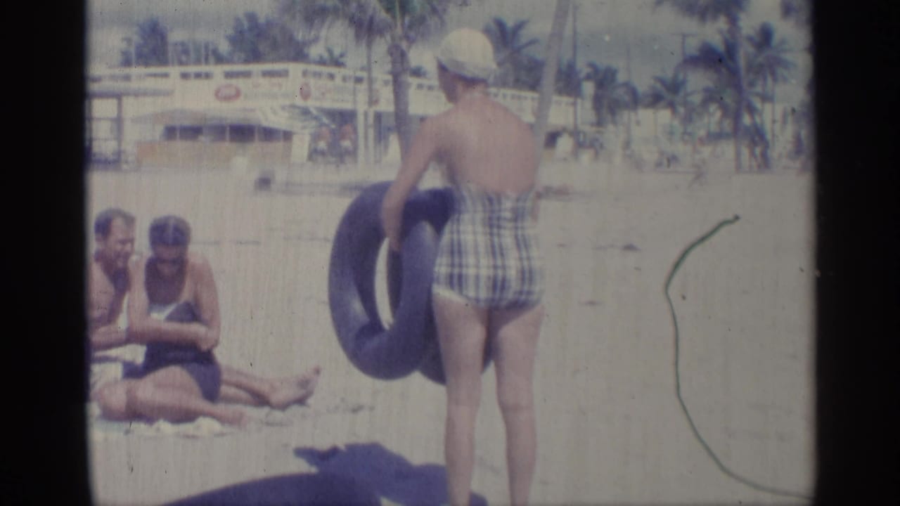 1959 Indiana home-movie frame showing a woman in a plaid swimsuit and white swim cap carrying a blue inner tube near beachgoers.