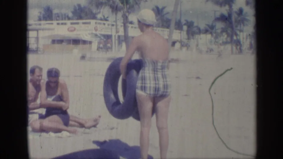 1959 Indiana home-movie frame showing a woman in a plaid swimsuit and white swim cap carrying a blue inner tube near beachgoers.