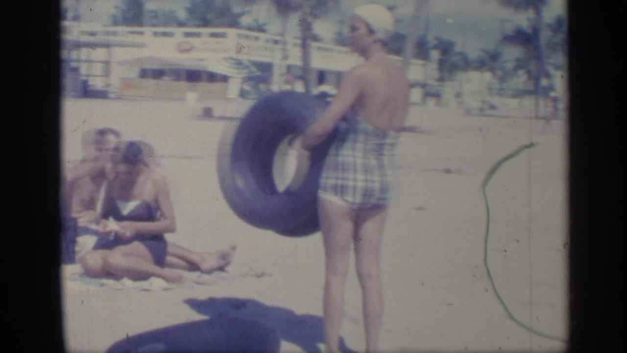 1959 Indiana beach scene with a woman holding a blue inner tube, seated adults on sand, and a palm-lined background building.