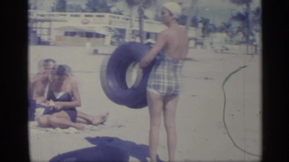 1959 Indiana beach scene with a woman holding a blue inner tube, seated adults on sand, and a palm-lined background building.