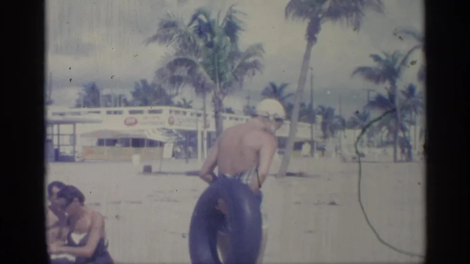 1959 Indiana home-movie frame of a woman in plaid swimwear bending near inner tubes, with seated beachgoers and palms in view.