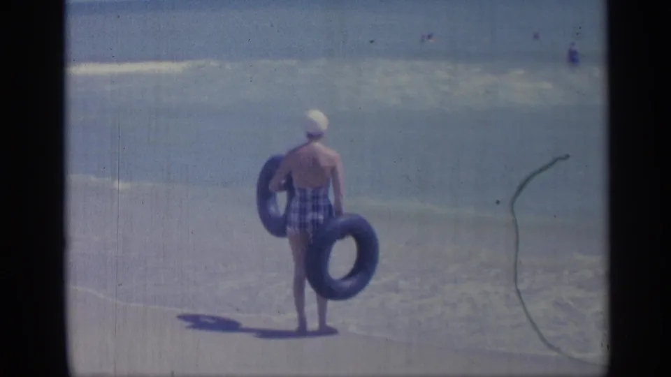 1959 Indiana beach frame showing a woman in a plaid swimsuit and white cap carrying two inner tubes toward the waterline.