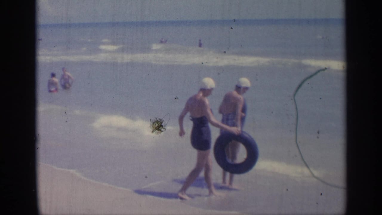 1959 Indiana 8mm frame of two women in one-piece swimsuits and white swim caps carrying inner tubes along a sandy shoreline.