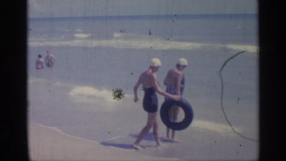 1959 Indiana 8mm frame of two women in one-piece swimsuits and white swim caps carrying inner tubes along a sandy shoreline.