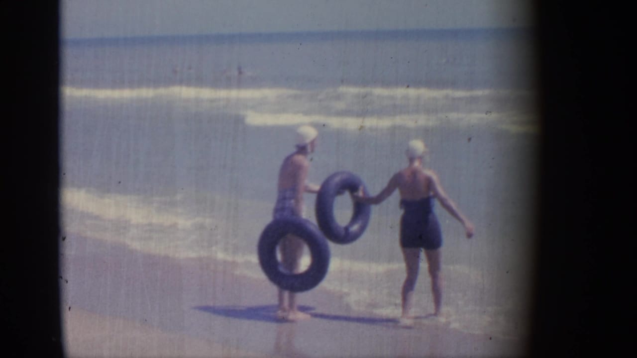 Two women with inner tubes stand at the shoreline near low waves.