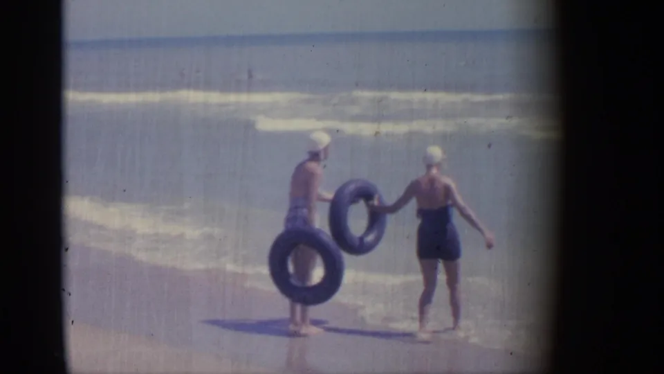 1959 Indiana beach footage showing two women in swim caps holding blue inner tubes at the edge of the water.
